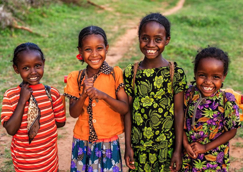 Four young girls in bright patterned dresses standing on a dirt path in rural Africa.