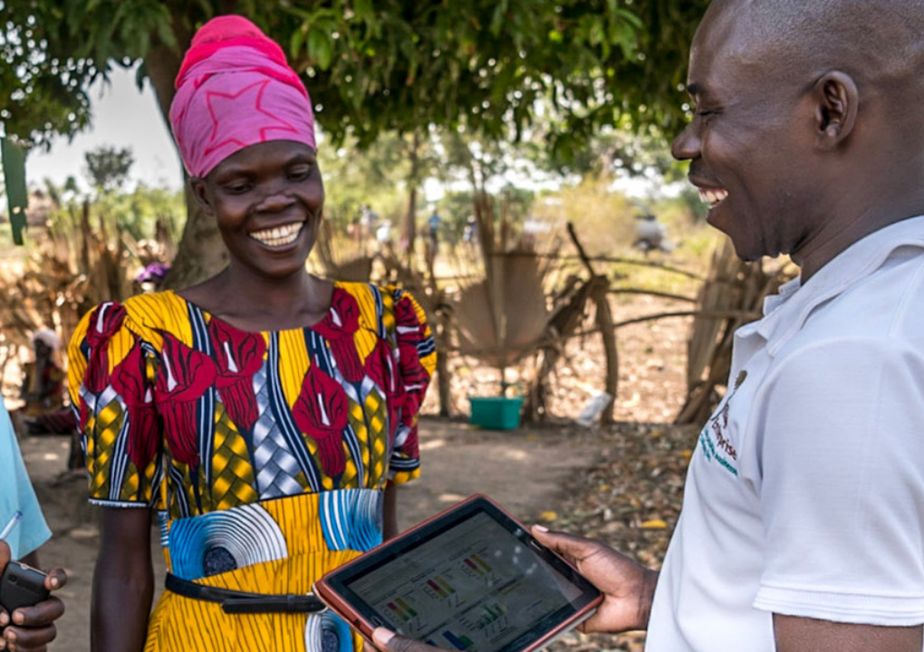 A Village Enterprise employee holding a tablet and laughing with a brightly dressed young woman in a village in Africa
