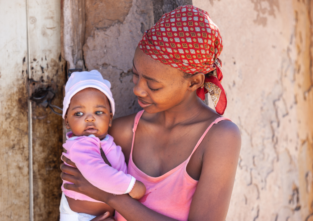 A young mom in a pink tank top and red head scarf smiling at her young daughter she's holding.