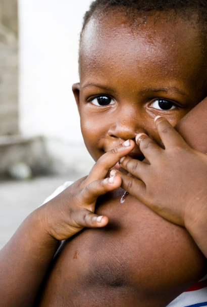 A young child looking into the camera, fingers raised to their lips